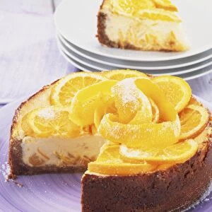 Baked St Clements cheesecake, topped with slices of orange and lemon and strips of orange and lemon zest, slice removed to show creamy inside of cake, stacked plates with slice on top in background