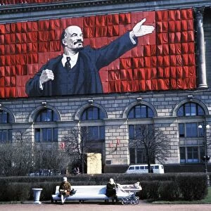 A banner of lenin on the side of a building overlooking two elderly women sitting on a park bench in leningrad, ussr, 1960s