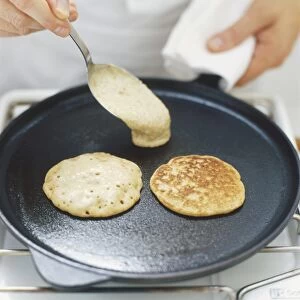 Blinis cooking on frying pan, raw mixture being added with a spoon, high angle view