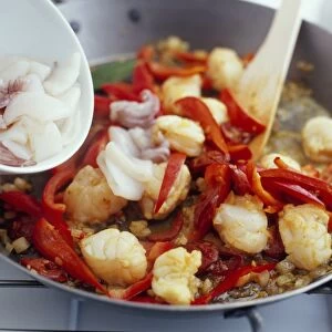Bowl of squid being added to a pan of prawns and red peppers to cook paella