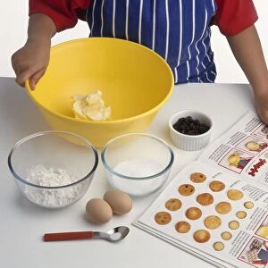 Boys hand pointing at ingredients in cooking book, mixing bowl containing butter, bowls of flour and sugar, eggs nearby