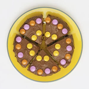 Chocolate cake decorated with colourful sugar-coated confectionery, cut into five pieces, served on yellow plate, view from above
