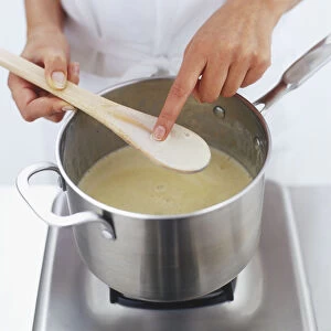 Finger being used to test the thickness of custard on wooden spoon over saucepan, high angle view