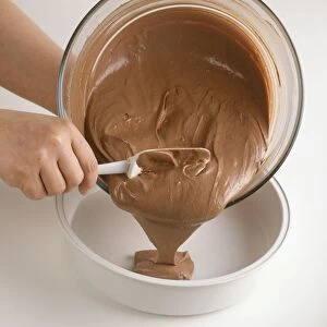 Hand holding mixing bowl and spoon, pouring chocolate cake mixture into cake tin