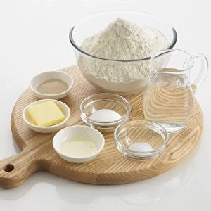 Ingredients required to make bread, including water, flour, salt, sugar, butter, yeast, in bowls and a jug, arranged on a chopping board