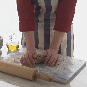 Kneading pasta dough on a marble board beside a rolling pin, jar of olive oil and a pestle and mortar