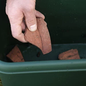 Mans hand placing broken clay pieces on bottom of green plastic trough, close-up