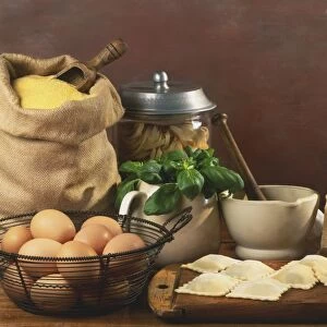 Pasta products and ingredients, including bag of durum wheat, jar of tagliatelle, block of parmesan cheese, bowl of eggs, pestle and mortar, cup of basil leaves, and fresh ravioli laid out on a chopping board