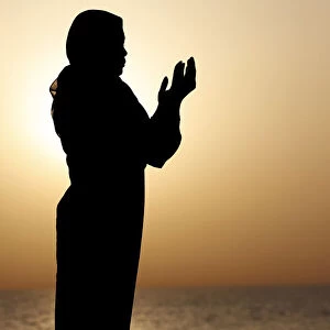 Silhouette of muslim woman in abaya praying with her hands up in air at sunset