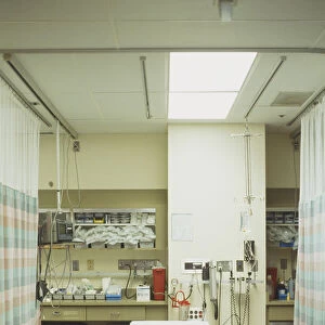Stretcher in hospital treatment area, sectioned off on either side by curtains, shelves for medical supplies and equipment in background