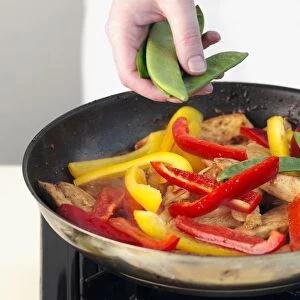 Womans hand adding mangetouts to chicken and peppers in frying pan, close-up