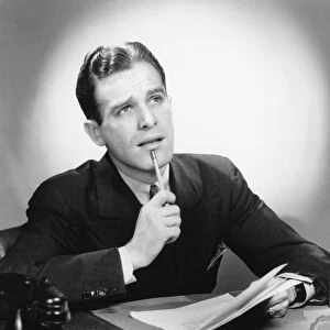 Man sitting at desk, holding pencil to lip, (B&W), portrait