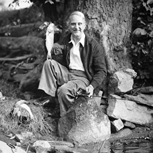 Senior man sitting by water, holding up fish (B&W), portrait
