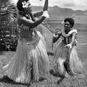 Two girls in grass skirts against a background of palms, flowers and mountains