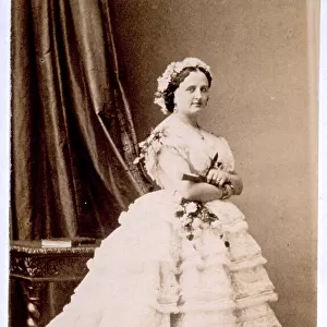 Portrait of a woman in evening dress with floral coiffure, holding a bouquet of flowers and closed fan. She is shown full-length, next to a table, on which a book is placed